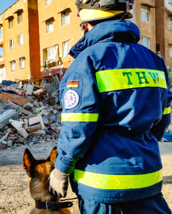 Rechts im Bild steht eine THW-Einsatzkraft mit einem Hund. Im Hintergrund liegen Trümmer eines eingestürzten Gebäudes.